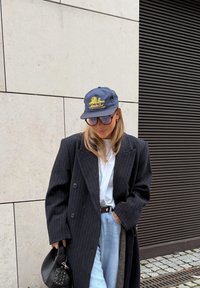 Long manteau noir à rayures, t-shirt blanc, jeans bleu clair, casquette marine avec design jaune, lunettes de soleil oversize, tenant un sac noir.