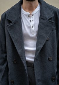 Cappotto grigio in lana doppiopetto con ampi rever, bottoni neri, indossato sopra una maglietta a coste bianca. Vestibilità su misura, tessuto testurizzato.