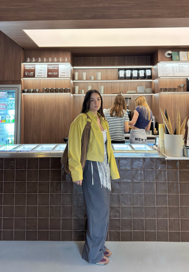 Jeune femme en veste jaune debout au comptoir d'un café moderne, avec deux personnes préparant des boissons derrière le comptoir.