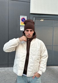 Weiße, gesteppte Bomberjacke mit Reißverschluss vorne, kombiniert mit einem braunen Hoodie. Das Modell trägt eine braune Mütze und hellblaue Jeans.