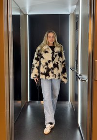 Fekete-fehér mintás műszőrme dzseki, kék farmer és fehér sportcipő, modern fekete és arany részletekkel díszített liftben állva.