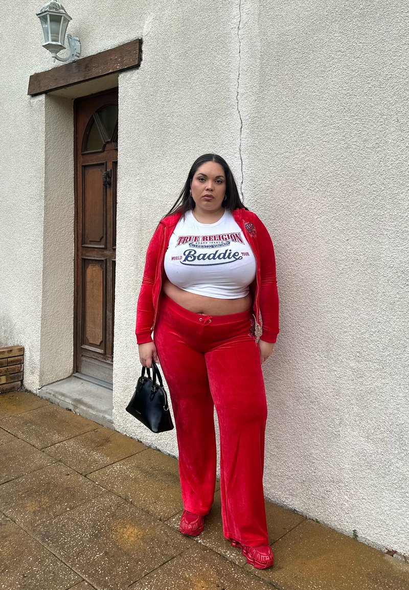 Femme debout à l'extérieur près d'un mur blanc texturé et d'une porte en bois, portant un survêtement rouge, un haut court blanc, tenant un petit sac à main noir.