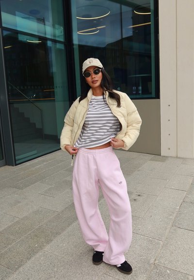 Mujer con chaqueta acolchada beige, camisa a rayas, pantalones deportivos rosa claro, gorra beige y gafas de sol, posando al aire libre junto a un edificio de cristal.