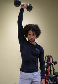 Black long-sleeve sports top, holding a dumbbell overhead. Background features various exercise weights and equipment.