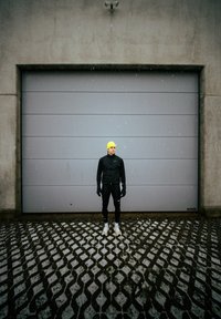 Homme en tenue de sport noire et casquette jaune fluo debout sur un trottoir à motifs légèrement enneigé devant une porte de garage grise fermée. Neige tombante.