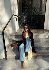 Cămașă maro peste un top nude, blugi evazați de culoare albastră deschisă și papuci bej. Ochelari de soare negri și o geantă maro așezate lângă. Trepte din piatră.