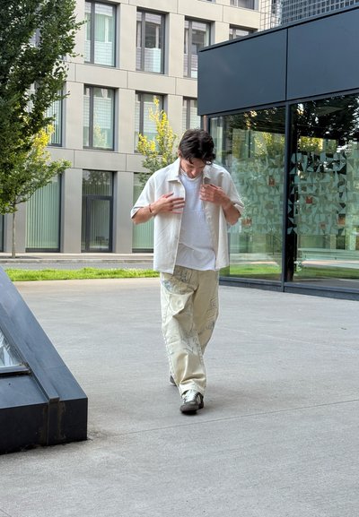 Una persona lleva una camisa de manga corta beige clara sobre una camiseta blanca, combinada con pantalones holgados beige con texto y diseños azules.