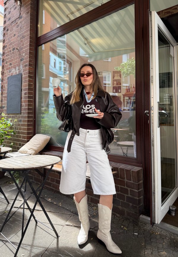 Femme avec des lunettes de soleil, une veste en cuir noire, un short blanc et des bottes blanches tient une tasse de café et une soucoupe devant un café un jour ensoleillé.