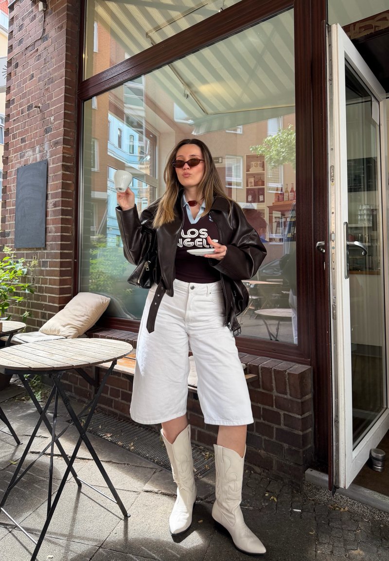 Femme avec des lunettes de soleil, une veste en cuir noire, un short blanc et des bottes blanches tient une tasse de café et une soucoupe devant un café un jour ensoleillé.