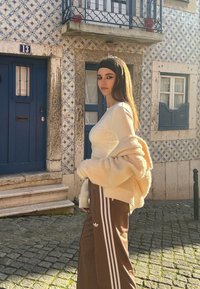 Krémový oversized sveter, hnedé tepláky s bielymi pruhmi, stojaci proti vzorovanej modro-bielej dlaždicovej stene s modrými dverami.