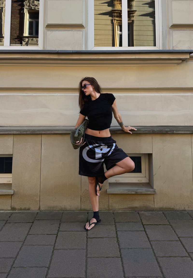 Maglietta nera aderente, pantaloni corti neri larghi con logo bianco e sandali neri. Sullo sfondo si vede una parete chiara e finestre.
