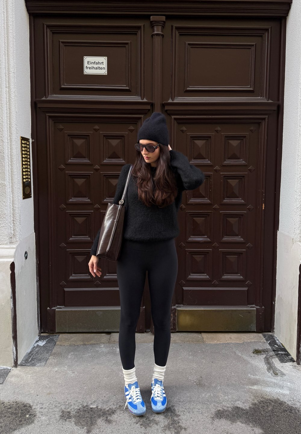 Črn puhast pulover, črne pajkice, modre superge z belimi vezalkami, krem nogavice. Rjava torba, oblečen v črno kapo in sončna očala, stoji ob vratih.