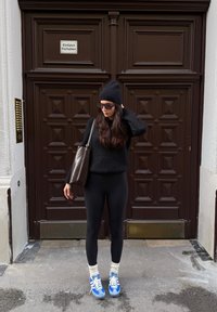 Črn puhast pulover, črne pajkice, modre superge z belimi vezalkami, krem nogavice. Rjava torba, oblečen v črno kapo in sončna očala, stoji ob vratih.