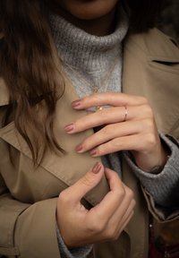 Casaco trench bege sobre um suéter cinza de gola alta; mãos com unhas curtas e ovais pintadas de rosa suave. Colar e anel delicados em ouro visíveis.