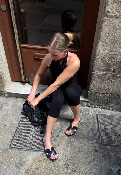 Bolso de cuero negro sobre una superficie de piedra. Persona con camiseta negra y leggings está sentada, lleva sandalias negras con un diseño geométrico recortado.