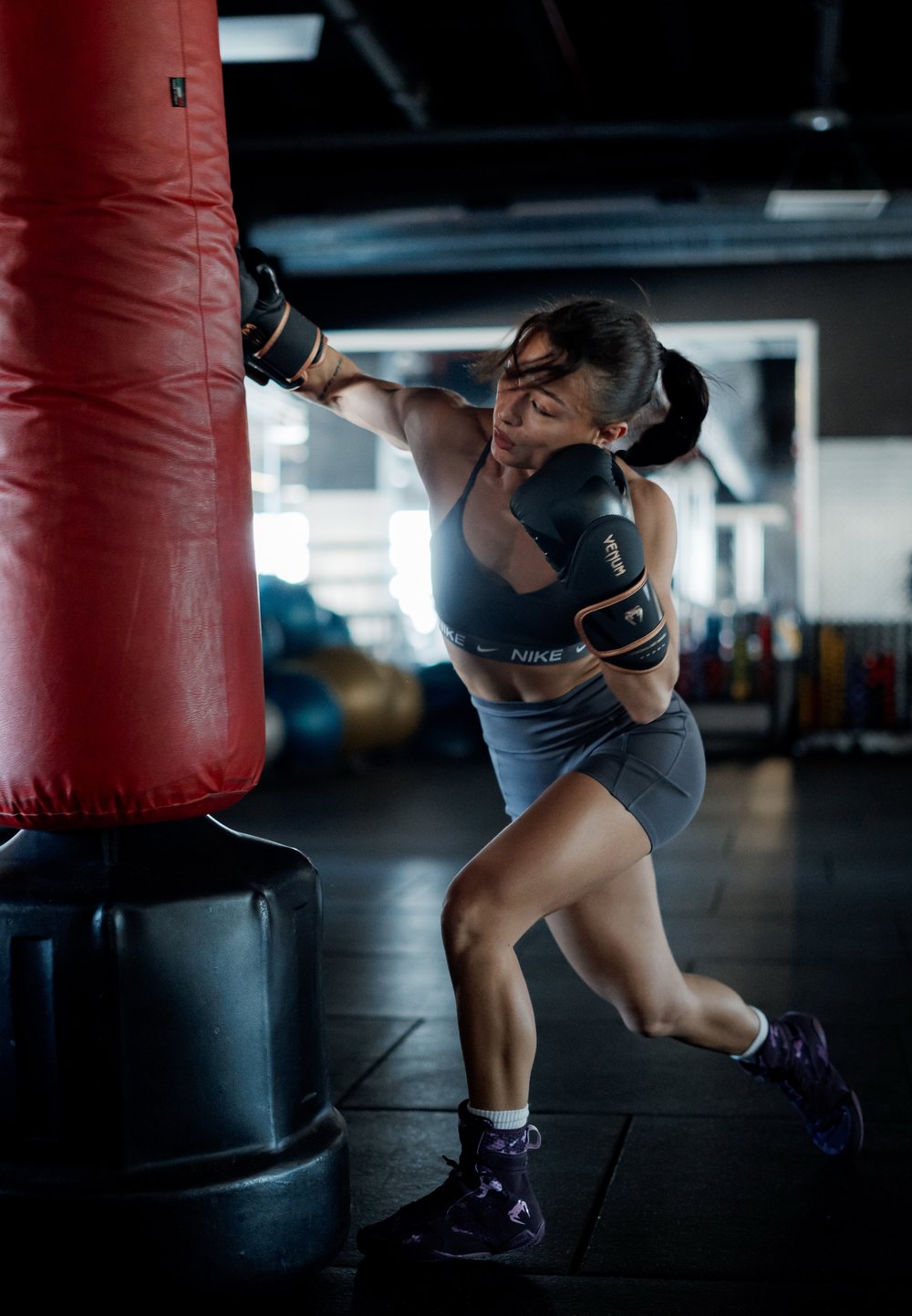Boxeuse en soutien-gorge de sport gris et short frappe un sac de frappe rouge. Gants noirs, chaussures foncées avec des accents violets. Environnement de salle de gym avec un sol sombre.