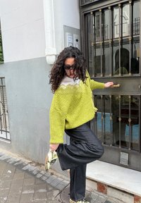 Femme en pull vert ajuste sa chaussure tout en tenant la poignée de la porte sur un trottoir urbain près d'un mur de bâtiment gris.