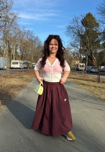 Mujer de pie en un camino iluminado por el sol, vestida con un top rosa y blanco, falda bordó de Nike, zapatillas amarillas y sosteniendo un bolso pequeño de neón amarillo.