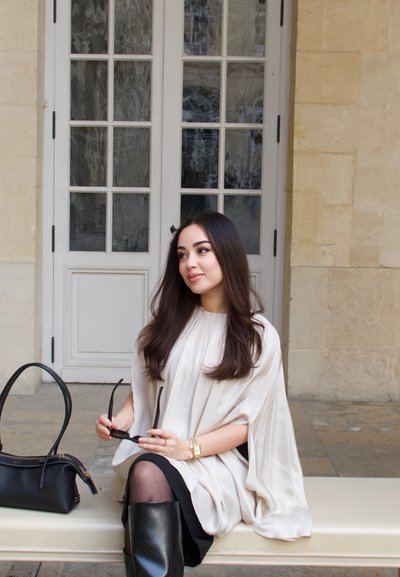 Joven mujer con cabello largo y oscuro, con blusa blanca y falda negra, sentada en un banco, sosteniendo gafas de sol, bolso negro a su lado, edificio beige de fondo.