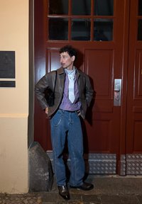 Person med lockigt hår i brun läderjacka, lila rutigt skjorta, vit bandana, blå jeans och mörka skor står mot en röd dörr på natten.