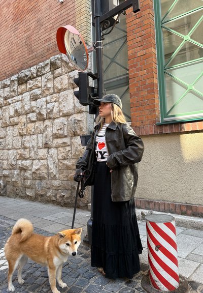 Mujer con chaqueta de cuero negra y falda larga de pie en una calle empedrada con un perro con correa cerca de un espejo de tráfico y un bolardo con franjas rojas.