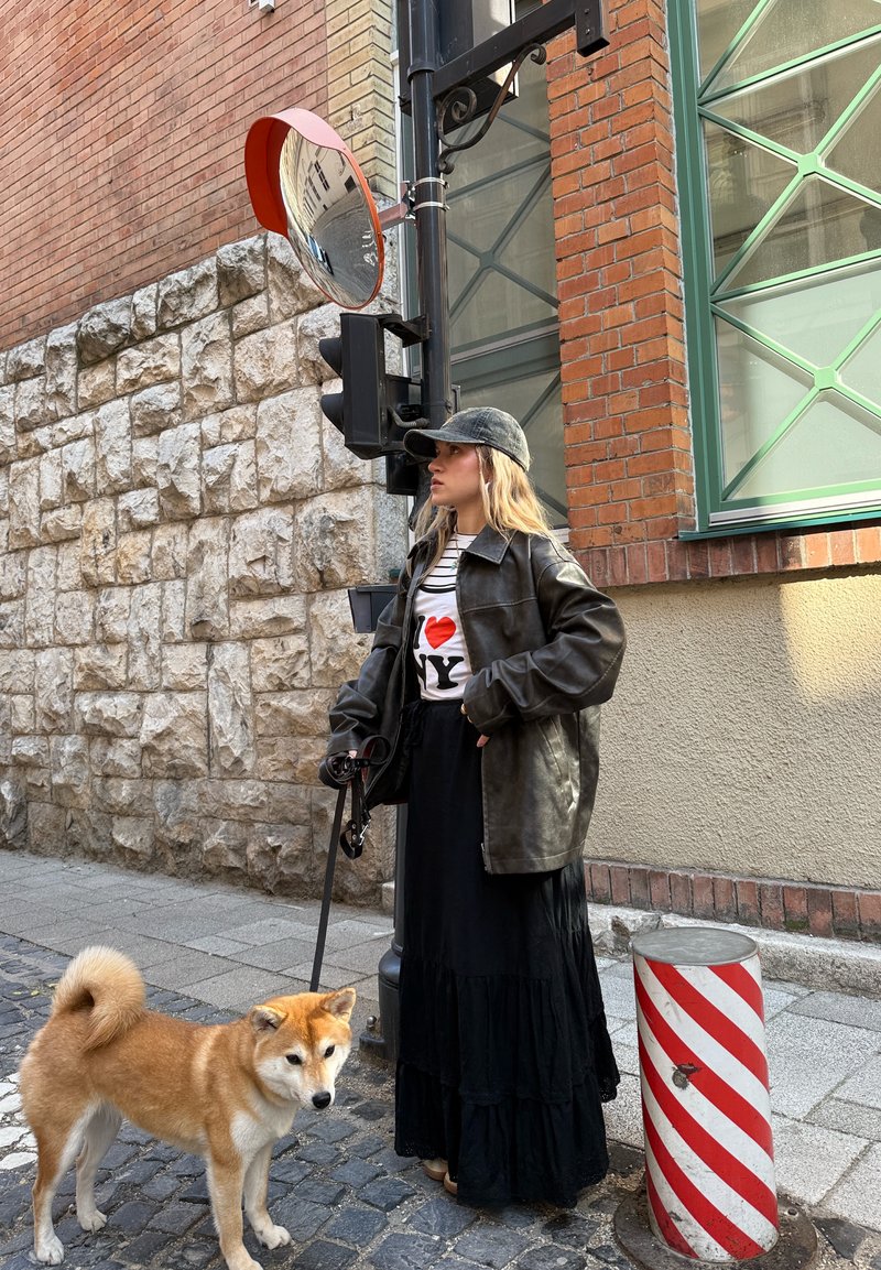 Femme en veste en cuir noire et longue jupe debout dans une rue pavée avec un chien en laisse près d'un miroir de circulation et d'un bollard rayé rouge.