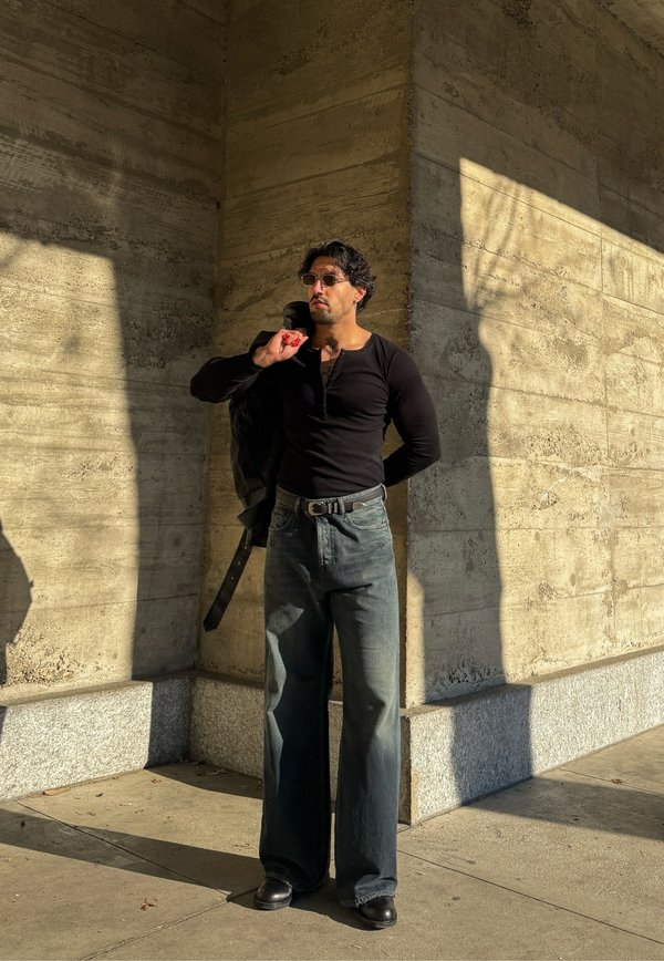 Άνδρας που φοράει μαύρο μακρυμάνικο μπλουζάκι και τζιν, κρατώντας το μπουφάν στον ώμο, στέκεται σε έντονο ηλιακό φως μπροστά σε μια υφή σκυρόδεμα.