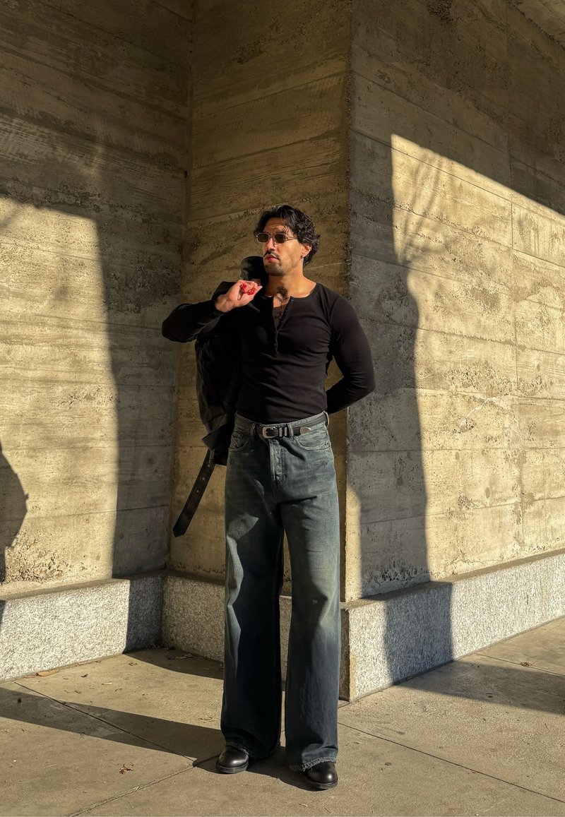 Man draagt een zwart shirt met lange mouwen en jeans, houdt een jas over zijn schouder, staat in fel zonlicht tegen een gestructureerde betonnen muur.