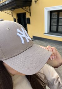 Gorra de béisbol beige con un logo bordado blanco. Presenta un visor curvado, costuras visibles y textura de tela.