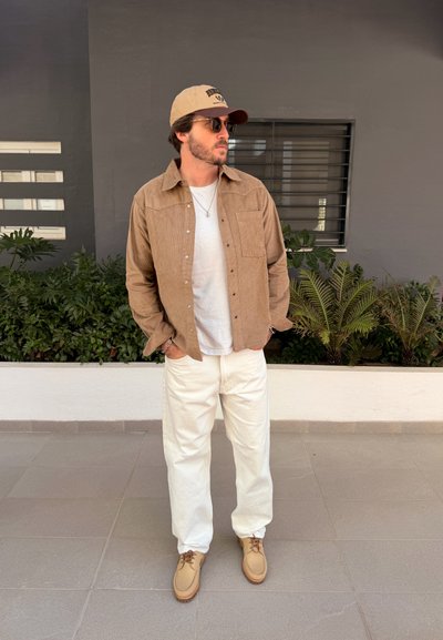 Hombre con gafas de sol, gorra beige, camisa marrón sobre camiseta blanca, pantalones blancos y zapatos beige, de pie con las manos en los bolsillos cerca de plantas.