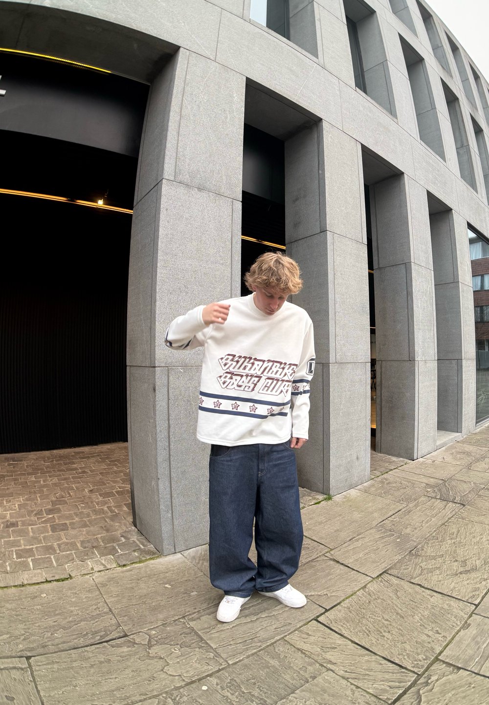 Λευκό πουκάμισο με μακριά μανίκια, γραφικά κείμενα και σχέδια αστεριών, συνδυασμένο με φαρδιά μπλε τζιν και λευκά αθλητικά παπούτσια. Αστική τοποθεσία με γκρίζα αρχιτεκτονική.