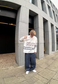 Λευκό πουκάμισο με μακριά μανίκια, γραφικά κείμενα και σχέδια αστεριών, συνδυασμένο με φαρδιά μπλε τζιν και λευκά αθλητικά παπούτσια. Αστική τοποθεσία με γκρίζα αρχιτεκτονική.