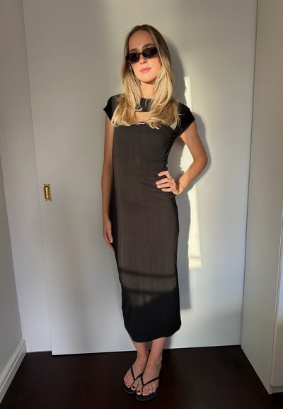 Mujer rubia vistiendo un vestido midi negro, chanclas negras y gafas de sol oscuras, de pie con una mano en la cadera frente a un fondo de puerta blanca.