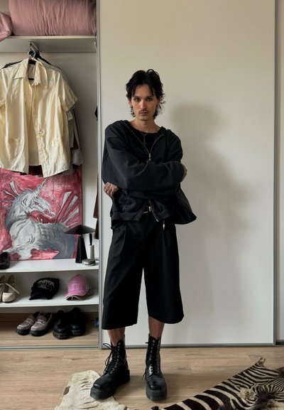 Persona con cabello oscuro vistiendo una sudadera negra de gran tamaño, pantalones cortos anchos negros y botas de plataforma negras, de pie con los brazos cruzados en una habitación con ropa y arte.
