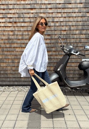 Weißes oversized Hemd, blaue Jeans, beiger geflochtener Einkaufstasche mit "MRS JEANS" in Hellblau. Der Hintergrund zeigt eine Holzwand und einen schwarzen Roller.