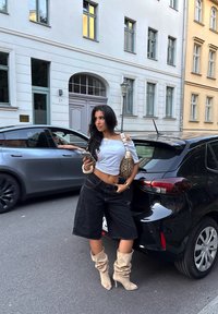 Pantaloni shorts neri oversize, top corto bianco, stivali beige con tacco alto e morbido, borsa con fantasia e telefono in mano, accanto a un'auto nera.