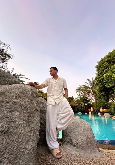 Camisa blanca de lino y pantalones sueltos con textura se destacan frente a una roca gris junto a una piscina azul. Fondo claro con vegetación y palmeras.