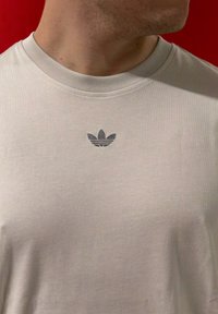 Jasnobeżowa koszulka z krótkim rękawem z okrągłym dekoltem i ciemnym, haftowanym logo na piersi. Gładka faktura bawełnianej tkaniny.