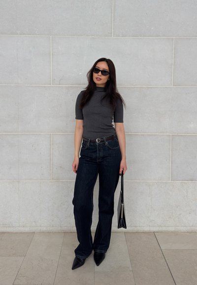Mujer con gafas de sol negras, camiseta de manga corta gris oscuro, jeans oscuros, zapatos puntiagudos y sosteniendo un bolso negro, de pie contra una pared clara.
