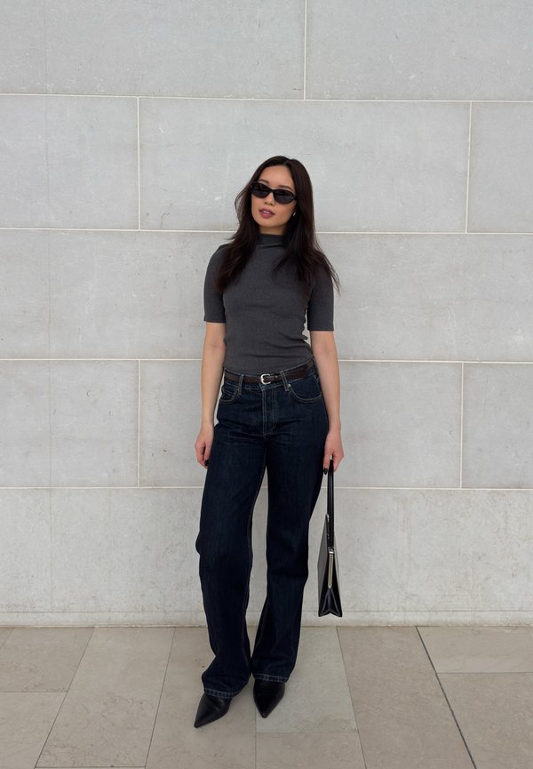 Vrouw met zwarte zonnebril, donkergrijze top met korte mouwen, donkere spijkerbroek, puntige schoenen en een zwarte handtas, staand tegen een lichte muur.