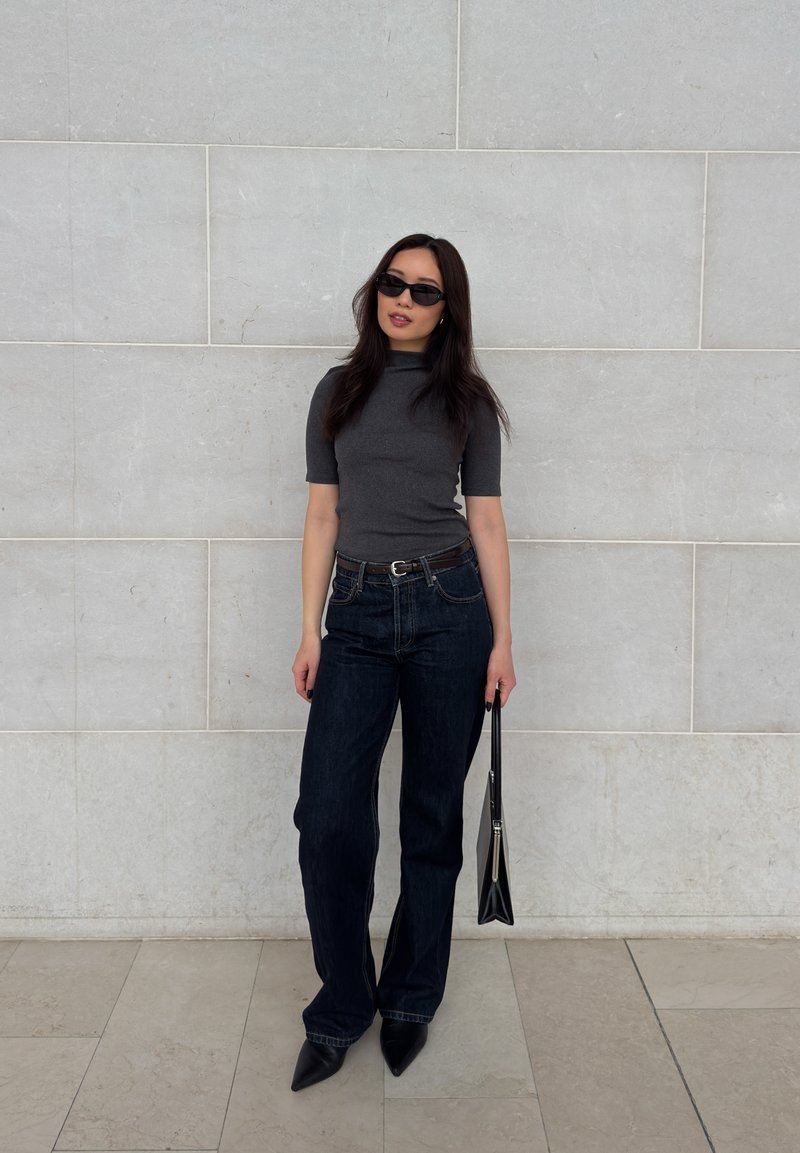 Γυναίκα με μαύρα γυαλιά ηλίου, σκούρο γκρι κοντομάνικη μπλούζα, σκούρο τζιν, μυτερά παπούτσια, που κρατά μαύρη τσάντα, στέκεται μπροστά σε έναν φωτεινό τοίχο.