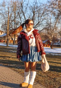 Rdeča usnjena jakna s ovitkom iz ovčje kože, bela kapuca z logotipom, kavbojka krilo, belknežne nogavice do kolen in rjavi polski čevlji. Osvetljen ozadje.