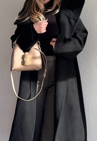 Kobieta w czarnym płaszczu i spodniach trzyma beżową torebkę z złotym zapięciem na tle jednolitej jasnej ściany.