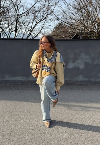 Mujer con gafas, camisa beige, bufanda azul a rayas, jeans claros y zapatos estampados levanta una pierna sobre una superficie pavimentada junto a una pared gris con árboles sin hojas.