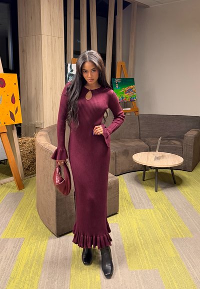 Mujer con vestido de canalé burdeos de manga larga con volante en el dobladillo y los puños, sosteniendo un bolso a juego, de pie en una habitación moderna con obras de arte y muebles.