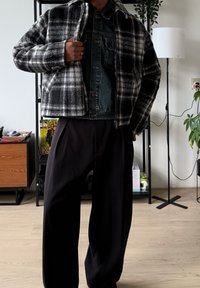 Veste courte à carreaux noir et blanc en laine, superposée sur une veste en denim. Pantalon plissé noir, éléments de fond minimalistes.