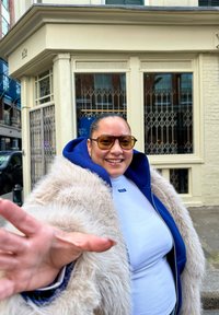 Une personne portant un haut blanc ajusté et un sweat à capuche bleu, recouverte d'un manteau en fourrure de couleur claire, avec de grandes lunettes de soleil jaunes, souriante.