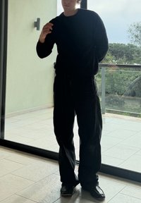 Νεαρό άτομο με κατσαρά μαλλιά που φοράει μαύρο πουλόβερ, φαρδιά μαύρη παντελόνια και μαύρα παπούτσια, στέκεται κοντά σε γυάλινη πόρτα με εμφανή εξωτερική βλάστηση.