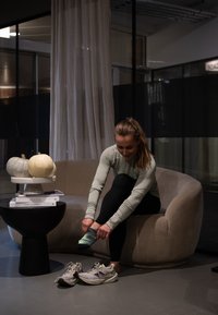 Moteris sėdi ant išlenktos sofos, užsidėdama sportines kojines šalia bėgimo batelių ir mažo staliuko su knygomis ir moliūgais prastai apšviestoje patalpoje.