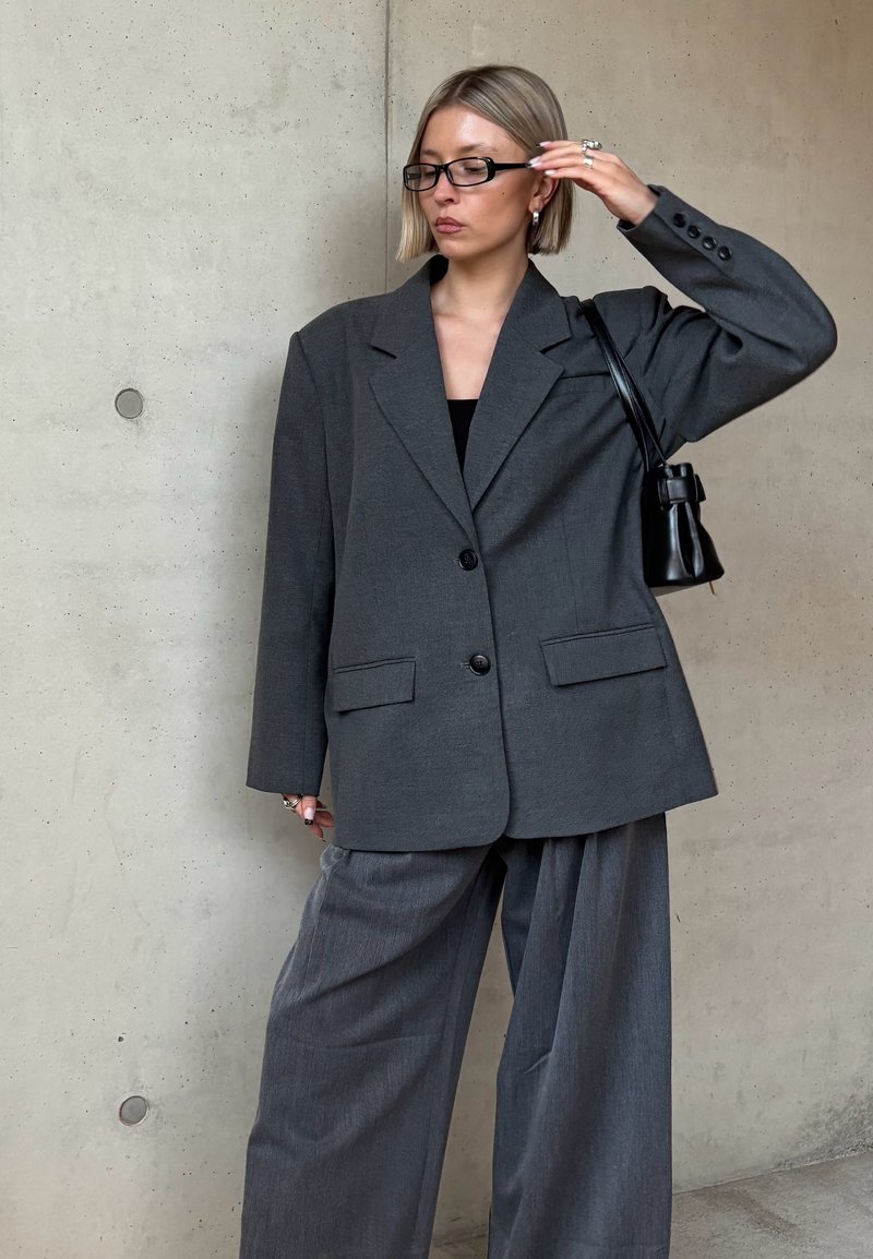 Woman with short blonde hair wearing black glasses, oversized dark gray blazer and wide-leg pants, holding black handbag against concrete wall.